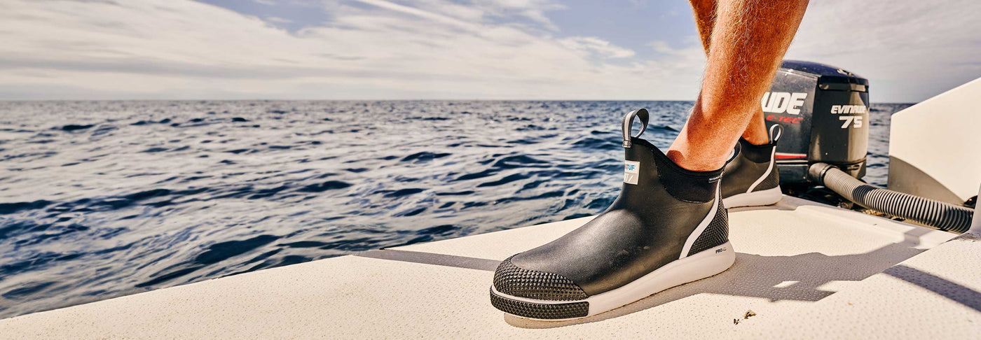 A close up photo of a man wearing a pair of black Xtratuf ADB Sport Boots on a motorised boat at sea