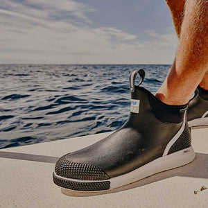 Close up of a man stood on the deck of a small motorised boat, wearing a pair of black Xtratuf ADB Sport Boots
