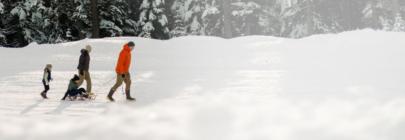 A family of four walking through the snow, wearing Xtratuf boots with the little boy on a sled being pulled by the father